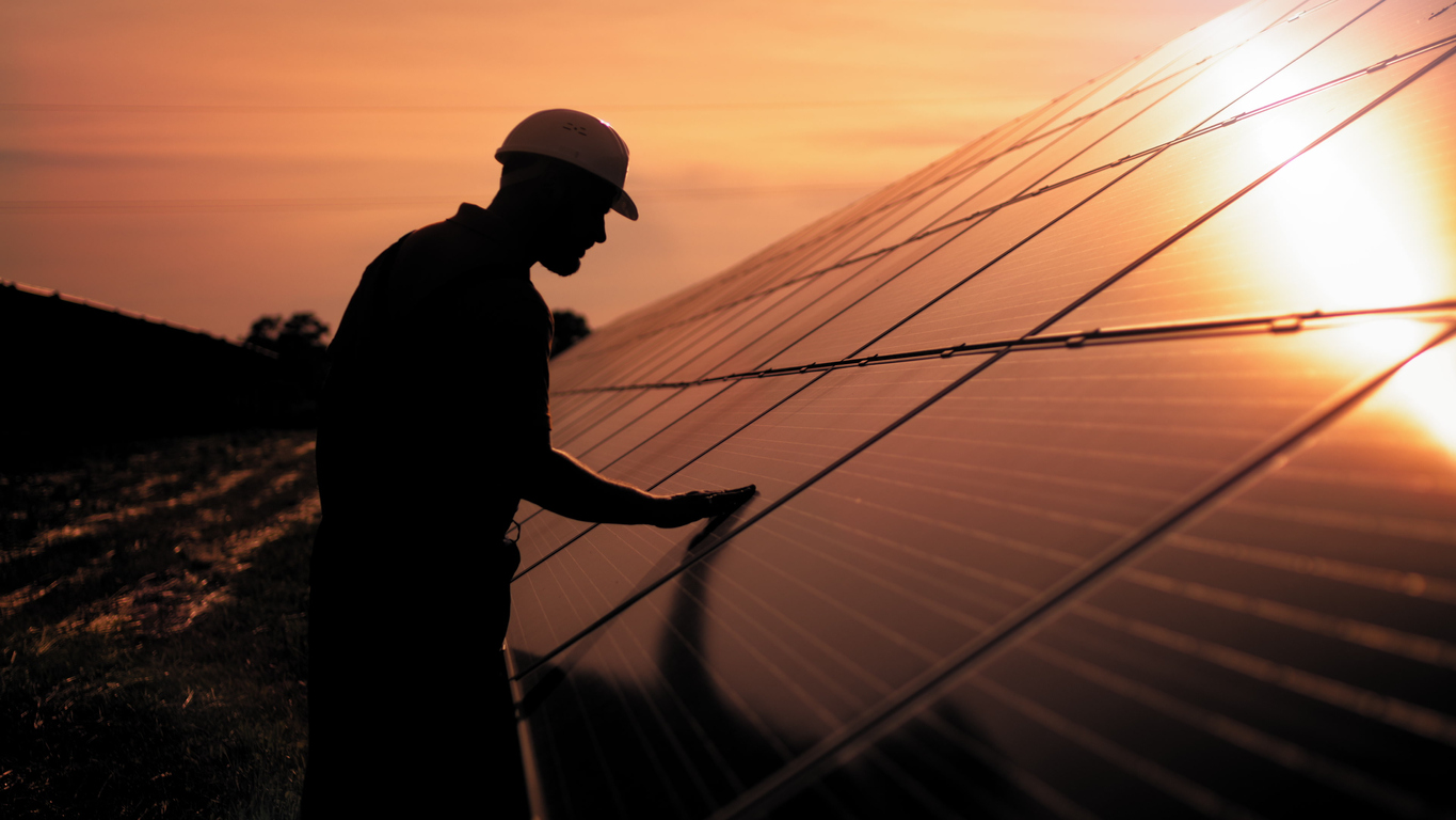 assistance technical worker in uniform is checking an operation and efficiency performance of photovoltaic solar panels. unidentified solar power engineer touches solar panels with his hand at sunset